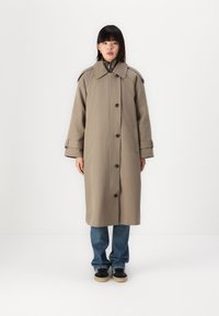 Abrigo largo beige de tela gruesa con un gran cuello, botones en el frente y bolsillos laterales. Lleva encima unos jeans azules y zapatos negros.