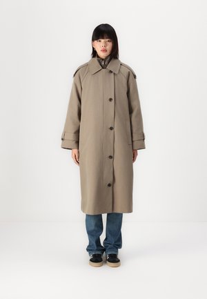 Beige lång kappa gjord av tjockt tyg med en stor krage, framknappar och sidofickor. Bärs över blåjeans och svarta skor.