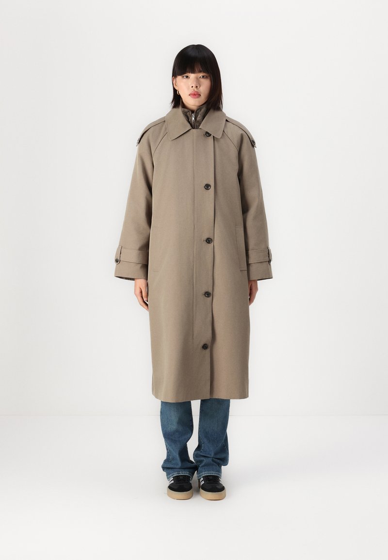 Abrigo largo beige de tela gruesa con un gran cuello, botones en el frente y bolsillos laterales. Lleva encima unos jeans azules y zapatos negros.