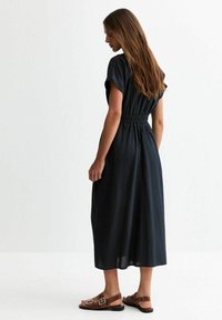 Mörkblå, medellång klänning med korta ärmar och elastisk midja. Tyget ser slätt och lätt ut, i kombination med bruna sandaler.