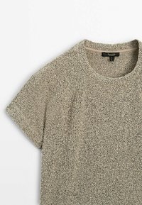 Haut en maille beige texturée à manches courtes avec une encolure ronde et une bande de couture beige visible à l’intérieur du col.