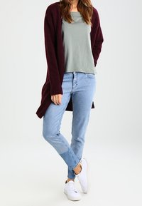 Bordeaux lang cardigan over en lys grøn top, parret med lyseblå jeans og hvide sneakers. Blød tekstur og afslappet pasform hele vejen igennem.