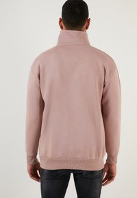 Sudadera rosa con cuello alto, puños y dobladillo acanalados. Textura suave, corte relajado y diseño minimalista. Sin gráficos o logotipos visibles.