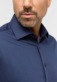 Camicia da uomo blu navy con colletto strutturato, texture liscia e un singolo bottone nero al colletto, presentata su uno sfondo chiaro.
