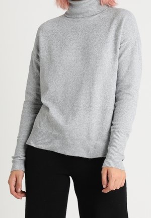 Maglione grigio a collo alto realizzato in un tessuto morbido, con polsini e orlo a coste, dalla vestibilità rilassata e una texture sottile. Indossato con pantaloni neri.