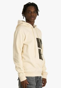 Felpa con cappuccio beige con tasca frontale di colore verde scuro. Presenta un cappuccio con coulisse, polsini a coste e una texture morbida.