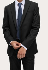 Completo nero con un design aderente, camicia bianca e cravatta in tessuto blu scuro. Stato aggiustando il polsino con il bordo bianco visibile.