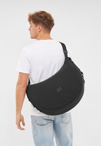 Černá crossbody taška s zaobleným tvarem a texturovanou látkou. Obsahuje nastavitelné ramenní popruhy a malou nášivku s logem na přední straně.