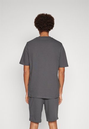 Mann mit kurzen lockigen Haaren, der ein einfaches dunkelgraues kurzärmeliges T-Shirt und passende Shorts trägt, mit dem Rücken zur Kamera.