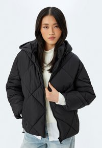 Sort pufferjakke med diamant quiltet mønster, stofhætte, lynlåslukning og elastiske manchetter, lagt over en hvid sweater.