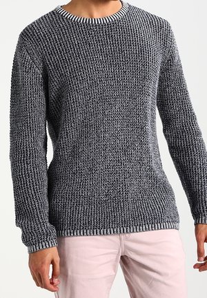 Man draagt een dikke grijze gebreide trui met een geribbeld patroon en een lichte broek, staande tegen een effen achtergrond.