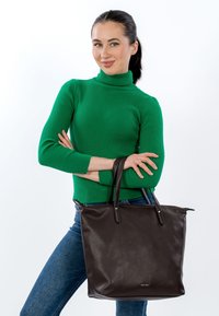 Rudos odos rankinė su tekstūruotu paviršiumi, turinti dvi ilgas rankenas ir viršutinį užtrauktuko uždarymą, laikoma žmogaus, dėvinčio žalią megztinį.