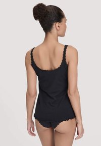 Vrouw met donker krullend haar in een knot, gekleed in een zwart topje met kant en bijpassend zwart ondergoed, met de rug naar ons gekeerd.