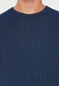 Pull en maille bleu marine avec un motif en câble texturé, présentant un col rond et un ourlet côtelé. Matière douce, design ajusté.