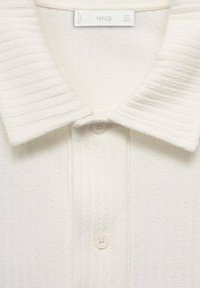 Camisa polo blanca hecha de tejido texturizado que presenta un cuello acanalado, tres botones y una etiqueta con "MNG" en el interior del cuello.