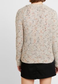 Pletený svetr v béžové barvě s multicolor náhodnými skvrnami, který má ribkovanou strukturu a uvolněný střih. Zadní strana má zaoblený lem a dlouhé rukávy.