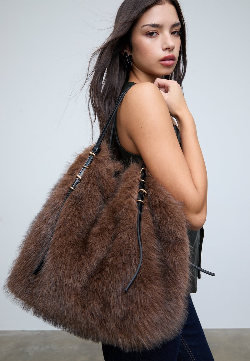 Brązowa torebka z futerka ekologicznego o miękkiej fakturze, czarnymi skórzanymi paskami z złotymi elementami oraz miękkim, zaokrąglonym kształtem.