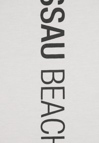 Hvidt stof med fed, sort tekst, der vertikalt skriver "NASSAU BEACH". Glat tekstur, enkelt design og tydelig skrift.