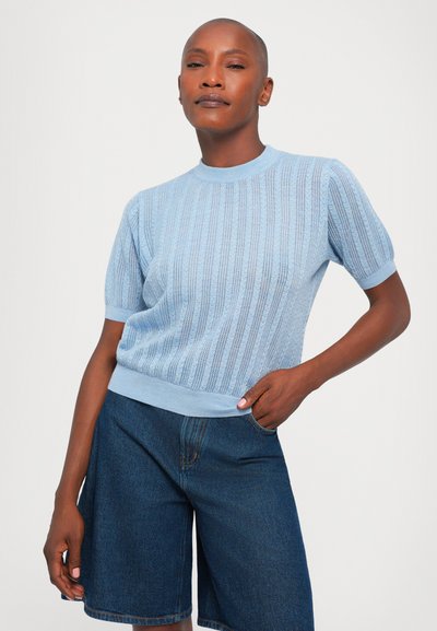 Pulover tricotat de culoare albastru deschis, cu mâneci scurte, design cu nervuri și guler rotund, asortat cu pantaloni scurți din denim albastru închis. Textură moale.