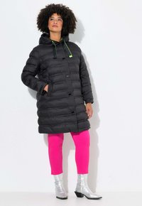 Abrigo de plumas negro con capucha, con acentos verdes y herrajes cuadrados. Lleva puesto encima de leggins rosas y botines plateados con cremallera.