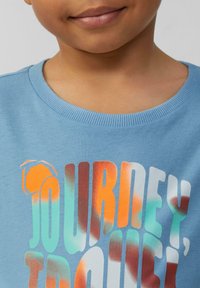 Lichtblauwe katoenen sweatshirt met gedurfde, veelkleurige tekst "JOURNEY, TROUBLE" met oranje, turquoise en witte accenten.