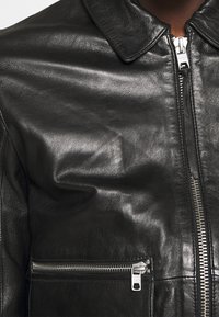 Svart läderjacka med en slät textur, krage och frontdragkedja. Inkluderar en ficka med dragkedja på vänster sida.