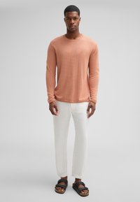 Camicia a maniche lunghe leggera e texturizzata color corallo, abbinata a pantaloni bianchi a gamba affusolata e sandali neri con design a cinturino.