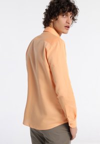 Chemise à manches longues orange clair en tissu lisse, avec un poignet à boutons et un col classique, présentant un ourlet légèrement incurvé à l'arrière.