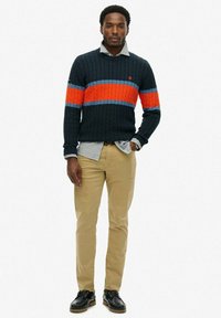 Marineblauwe kabeltrui met oranje en blauwe strepen, gecombineerd met een lichtblauw overhemd met kraag, beige broek en zwarte schoenen.