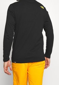 Eine Person trägt ein schwarzes Langarmshirt mit einem gelben Logo auf der hinteren Schulter und leuchtend orangenen Hosen, von hinten gezeigt.
