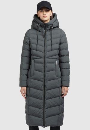 Woman wearing a long, dark gray quilted puffer coat with a hood and a black baseball cap, standing facing forward.