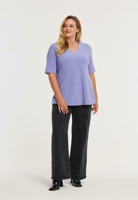 Lys lilla V-hals top lavet af blødt stof, parret med sorte plateaufede jeans med guld udsmykninger. Modellen står i en afslappet stilling.