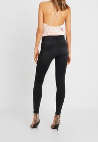 Mujer vestida con un camisón de satén rosa pálido, jeans ajustados negros de talle alto y sandalias de tacón alto de serpiente, de pie contra un fondo liso.