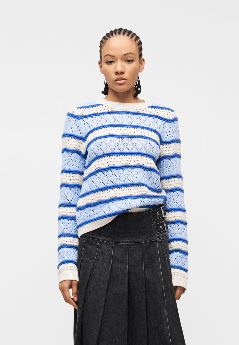 Νεαρό άτομο με πλεγμένα μαλλιά που φοράει πουλόβερ πλεκτό με ρίγες σε μπλε και κρεμ χρώμα και σκούρα πλισέ φούστα με πλευρικές αγκράφες.