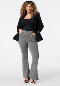 Blazer noir sur un haut noir ajusté, associé à un pantalon évasé gris avec des poches latérales et un tissu texturé. Des sandales à talons noires complètent le look.