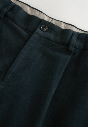 Mørkeblå bukser i blødt stof har flad front, en enkelt sort knaplukning og en kontrasterende beige foring indeni taljebåndet.