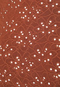 Tissu brun avec un motif en dentelle présentant des motifs floraux découpés. Surface texturée avec des accents blancs contrastants créés par les découpes.