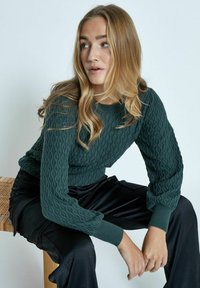 Maglione verde in maglia testurizzata con motivo a onda; polsini e scollatura a coste. Indossato con pantaloni in tessuto nero lucido.