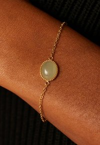 Gouden kettingarmband met een ovale groene steen, voorzien van een gestructureerd oppervlak en een parelrand, rustend op een gestructureerde huidtint.