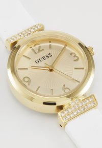 Une montre analogique dorée avec un bracelet en silicone blanc, présentant un cadran doré texturé, des chiffres noirs et des accents en cristal sur les cornes.