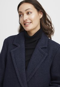Námornícky kabát z vlnenej zmesi s houndstooth vzorom, so špicatými klopami a voľným strihom. Nosený cez čierny rolák s textúrovaným povrchom.