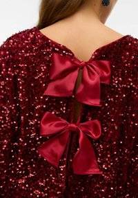 Rød top med palietter og stor åben ryg, fastgjort med tre satin sløjfer. Struktureret, skinnende overflade giver visuelt liv.