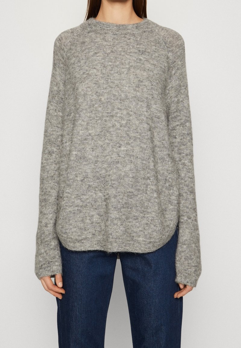 Femme portant un pull ample gris clair duveteux à manches longues et un jean bleu foncé, debout devant un fond clair uni.