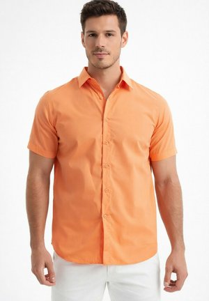 Homme portant une chemise à manches courtes orange vif boutonnée et un pantalon blanc, debout devant un fond blanc uni.
