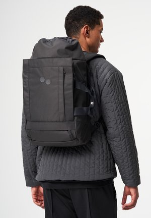 Man wearing dark quilted jacket carrying large black backpack with top flap and front zip pocket, standing against light background.