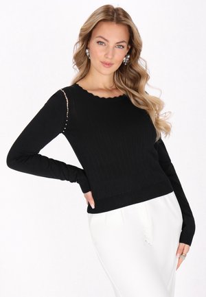 Vrouw met lang golvend haar, gekleed in een zwarte top met lange mouwen met pareldetails en witte broek, poseert met één hand op de heup tegen een witte achtergrond.
