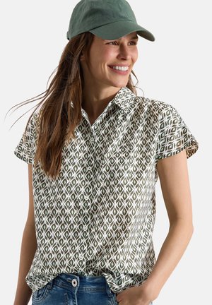 Uśmiechnięta kobieta w zielonej koszuli z krótkim rękawem w wzory, niebieskich dżinsach i zielonej czapce z daszkiem, z długimi brązowymi włosami.