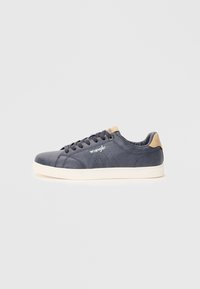 Baskets en cuir bleu marine avec des perforations, semelle en caoutchouc blanche, accent de talon marron et logo "Wrangler" sur le côté.