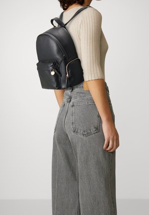 Svart ryggsäck i läder med rundad form, försedd med en framficka med dragkedja, detaljer i guldfärg och justerbara remmar. Stylad med grå denimbyxor.