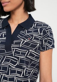 Γυναίκα φοράει μπλε ναυτικό πόλο με λευκές αφηρημένες γραμμές και μικρό κόκκινο, λευκό και μπλε λογότυπο στο στήθος.